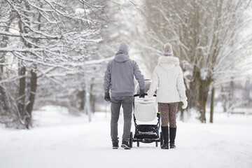 Young family pushing white baby stroller and walking on snow covered sidewalk at park in cold winter day. Enjoying peaceful stroll. Spending time with newborn and breathing fresh air. Back view. - obrazy, fototapety, plakaty