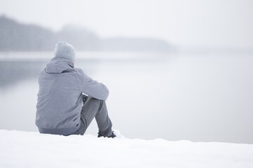 Young adult man sitting alone on snow at lake shore and looking far away. White cold snowy winter...