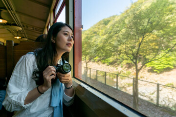 curious asian Japanese female visitor taking Arashiyama Mountains sightseeing train in Kyoto japan. she cranes neck to look out of window while trying to take picture with slr camera