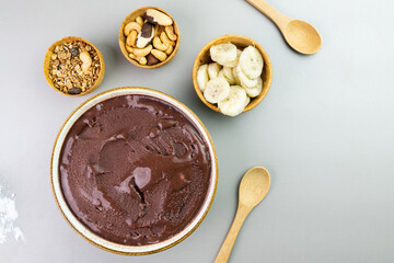 Brazilian açai in a white bowl, additional banana, granola and nuts in small bamboo bowls. Cashew and Pará nuts. Top view. Gray background. Selective focus