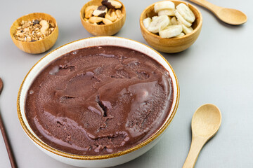 Brazilian açai in a white bowl, additional banana, granola and nuts in small bamboo bowls. Cashew and Pará nuts. Top view. Gray background. Selective focus