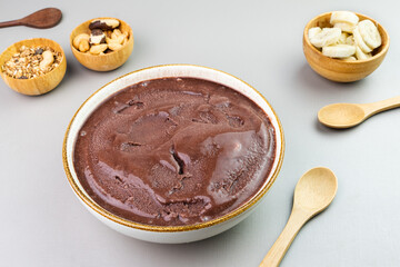 Brazilian açai in a white bowl, additional banana, granola and nuts in small bamboo bowls. Cashew and Pará nuts. Top view. Gray background. Selective focus