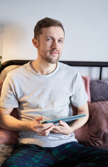 Cheerful bearded male at home working using tablet computer. Young handsome man in gray t-shirt sitting on bed making online shopping or surfing the internet. High quality vertical image