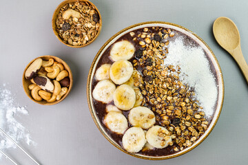 Brazilian açai in a white bowl with banana, powdered milk, honey, granola and nuts in small bamboo bowls. Cashew nuts and Pará. Top view. Grey background. Selective focus