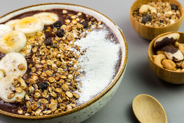 Brazilian açai in a white bowl with banana, powdered milk, honey, granola and nuts in small bamboo bowls. Cashew nuts and Pará. Grey background. Selective focus