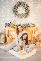 Young woman with child baby girl 7 years old in sleepwear watching film and eating popcorn on the bed in Christmas decorated home