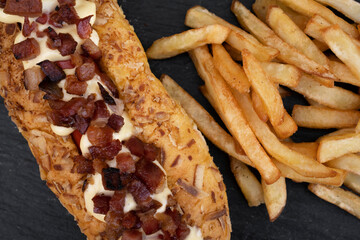 Hot dog with crispy bacon, alioli sauce and french fries in a black dish.