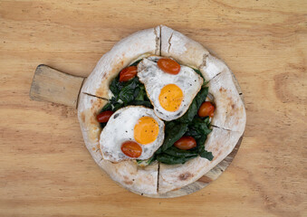 Top view of a pizza with mozzarella cheese, spinach, cherry tomatoes and fried eggs, on the wooden table.
