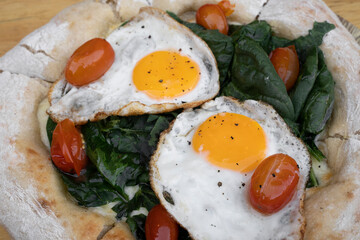 Closeup view of a pizza with mozzarella cheese, spinach, cherry tomatoes and fried eggs, on the wooden table.