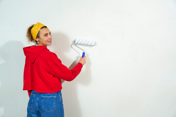 A young female artist paints the walls in the apartment with a roller. Beautiful woman is working on a new bedroom project. Cute woman in blue jeans and red hoodie painting a wall. Back view.