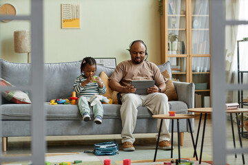 Full length portrait of black single father with cute son sitting on couch in messy home interior, man working with baby, copy space