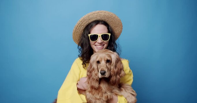 Stylish woman with dog in summer. Static shot of happy young female owner in stylish sunglasses and straw hat caressing and kissing English Cocker Spaniel dog against blue background.