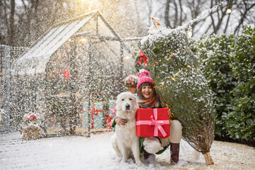 Portrait of a woman with Christmas tree and giftbox hugs with her adorable dog at beautifully decorated yard on snowfall. Concept of happy winter holidays and magic