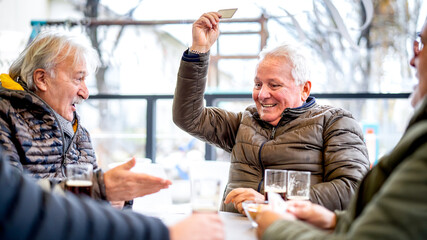 Senior friends playing cards at local bar on winter day - Ageless lifestyle concept with mature...