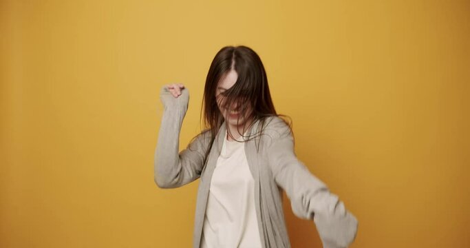 Happy young brunette dancing energetically. Merry young woman in stylish clothes shaking long dark hair and smiling gleefully while performing energetic dance against yellow background