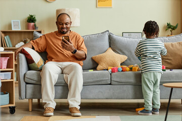 Full length portrait of father using smartphone with son playing on sofa at home, copy space