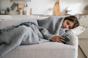 Upset sad teenage girl lying under blanket on sofa holding phone, kid spending time staring at...