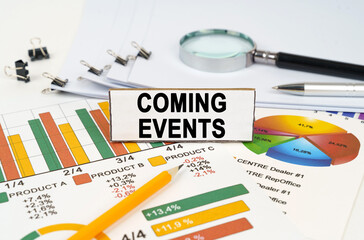 On the business charts lies a magnifying glass, a pen and a sign with the inscription - Coming Events