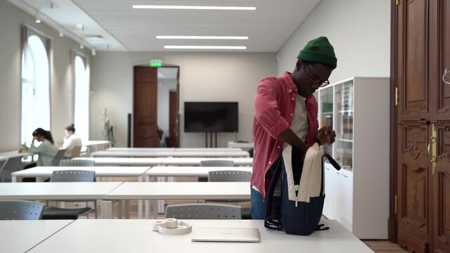 Pedantic African American male student laying out things on desktop in library to prepare for college exams or study. Composed black man pushes back chair sits at seat opens laptop for work on project