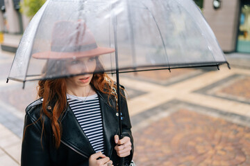 Shooting through transparent umbrella to elegant redhead young woman in fashion hat standing on...