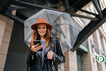 Low-angle view of cheerful elegant young woman wearing fashion hat using typing mobile phone smiling looking to screen standing with transparent umbrella, by building in rainy autumn city street.