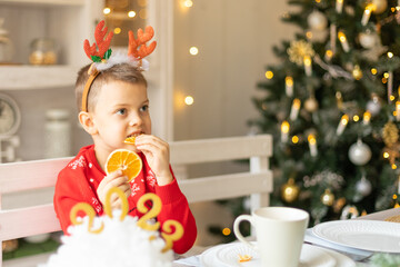 Cheerful little boy wearing warm red xmas sweater, deer horns on head eat dry orange slices in the Christmas decorated kitchen and drinking hot chocolate. Fresh healthy food. Happy New Year concept