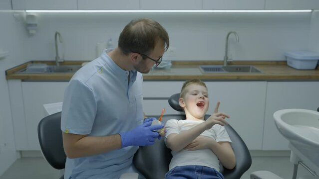 Boy with his mouth open sits in dental chair and shows his teeth to dentist. Doctor has toothbrush and artificial jaw in his hands. Slow motion
