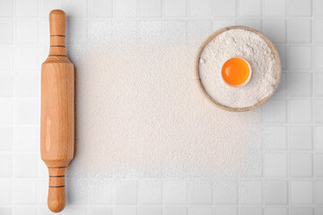 Flour, rolling pin and raw egg on white tiled table, top view