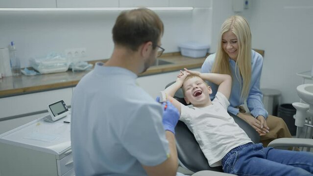 Professional male dentist shows boy and his mother how to brush their teeth in playful way on model of jaw. Child sitting in dental chair and laughing. Mom smiles. Slow motion