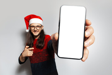 Studio portrait of young happiness man, holding big smartphone with blank on screen in hand, pointing at device, wearing Christmas sweater of red color and Santa hat, on a white background.