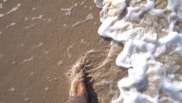 Barefooted man walking along the beach with waves crashing against his feet in slow motion