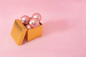 A golden gift box with pastel pink Christmas baubles on pink background. Minimal pastel concept for season greetings card or banner. Design for New Year 2023 celebration party invitation. Copy space