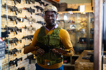 African-american man wearing armor vest and helmet while standing with machine gun in airsoft shop.