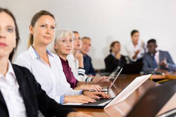 Fototapeta premium Professional middle-aged white blond businesswoman sitting among colleagues at conference table in meeting room and listening to speaker's presentation