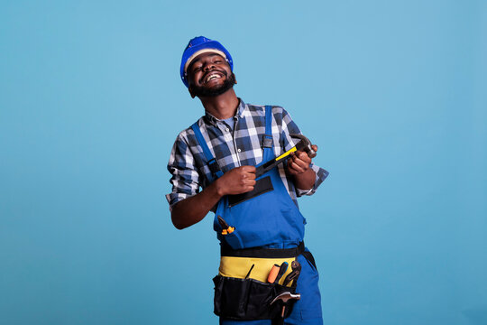 Studio Photo Of Glad African American Builder In Helmet And Utility Belt Holding Hammer With Two Hands. Happy Construction Worker Smiling Proudly On Blue Background In Studio Shot.