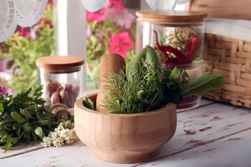 Mortar with pestle and fresh green herbs on white wooden table near window
