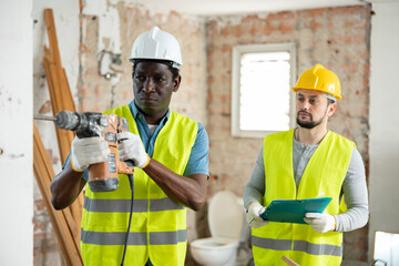 African american man construction worker using demolition hammer. Caucasian man standing behind and checking documentation.