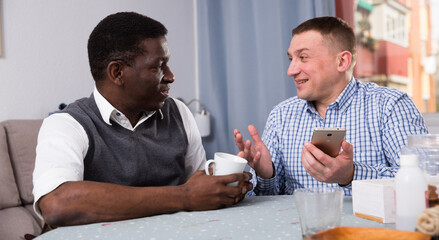 Interracial two male friends using phone and talking in home interior