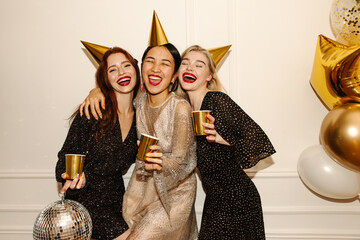 Three joyful young interracial women having drinking party on white background. Beauty wearing sparkly dresses laugh with their eyes closed. Holiday concept