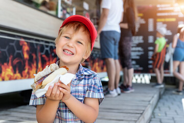 Cute adorable little happy smiling boy kid enjoy eating hot dog sausage in bread near street cafe stall outdoors. Child healthy eating lunch hotdog. Junk food and fast food unhealthy snack concept