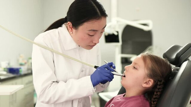 Cute pretty little girl during the dental procedure at the modern children's dental clinic. Children's asian female dentist curing teeth of a girl, using dental drill and mirror.