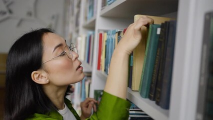Asian woman looks through titles of books on library shelf