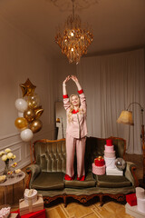 Attractive young caucasian girl wearing pajamas is standing on couch with hands up in room. Blonde celebrates her birthday in decorated room. Emotion concept