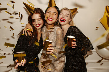 Three elegant young interracial girls at night party are having fun along with booze and confetti. Blondes, brunettes and redhead wear sparkly dresses. Birthday concept