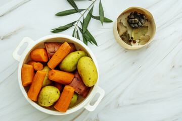 A white plastic container filled with cleaned and chopped potatoes, sweet potatoes and carrots ready for cooking on a white marble surface with dried leaves - Powered by Adobe