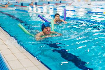 Active positive caucasian married senior adult couple swimming in a sport pool. People in swimming caps using colourful foam pool noodles for support. High quality photo