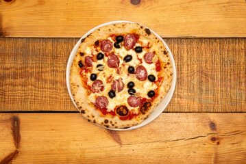 A Neapolitan pizza with black olives, pepperoni, mozzarella and grated Parmesan cheese and tomato cooked in a stone oven on a wooden table