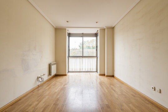 An empty living room with oak type wood laminate flooring and soft cream colored walls in need of a fresh coat of paint
