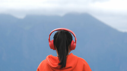 Rear view of a young woman standing on a calm hilltop and listening to music in headphones in the morning. Woman wearing a sweater enjoying the beauty of nature looking at the mountain in winter.