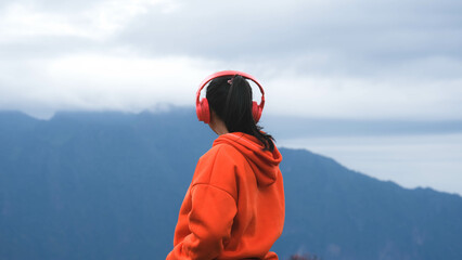 Rear view of a young woman standing on a calm hilltop and listening to music in headphones in the morning. Woman wearing a sweater enjoying the beauty of nature looking at the mountain in winter.